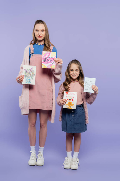 full length of mother and happy daughter holding greeting cards on purple, mothers day concept - Фото, изображение