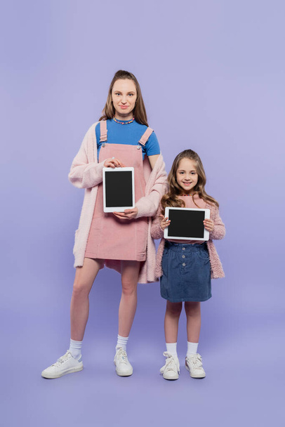 longitud completa de madre e hija sosteniendo tabletas digitales con pantalla en blanco en púrpura - Foto, Imagen
