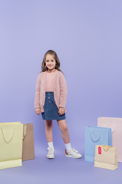 full length of happy kid standing near shopping bags on purple - Foto, immagini