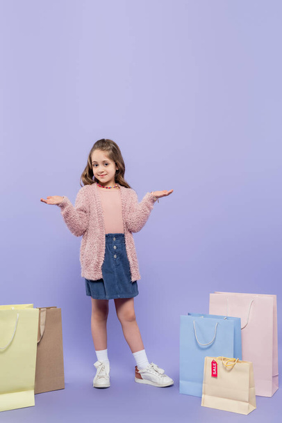 full length of happy kid pointing with hands near shopping bags on purple - Foto, afbeelding