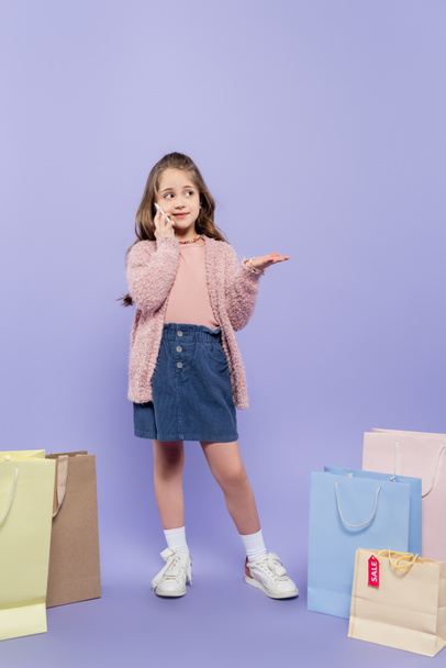 full length of happy kid talking on smartphone and standing near shopping bags on purple - Foto, immagini