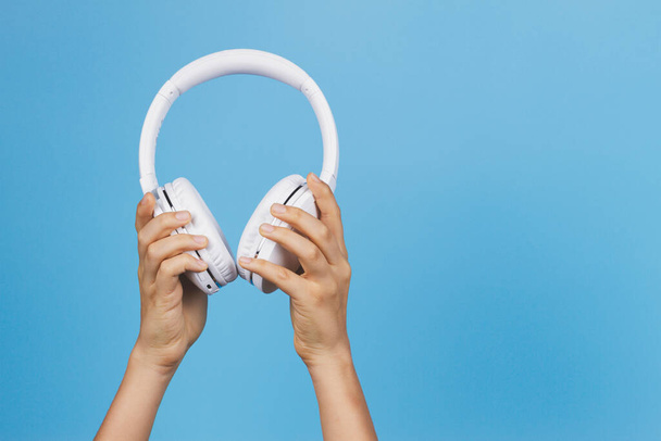 Raised hands holding modern white wireless headphones on light blue background. Front view - Foto, Imagem