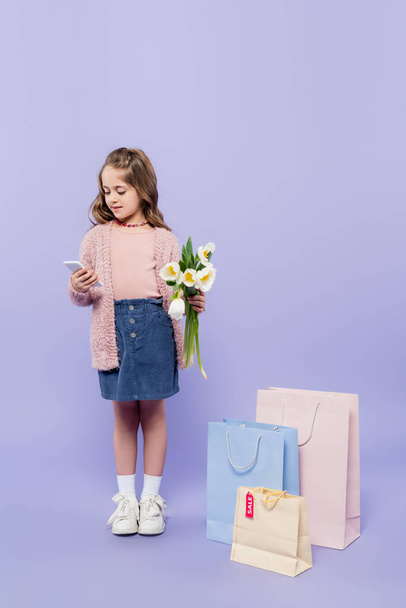 full length of kid holding flowers and using smartphone while standing near shopping bags on purple - Fotó, kép