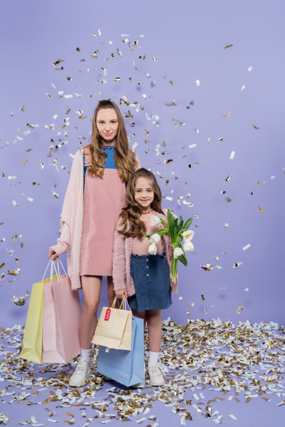 in voller Länge glückliche Mutter und Tochter mit Einkaufstaschen und Blumen neben fallendem Konfetti auf lila - Foto, Bild