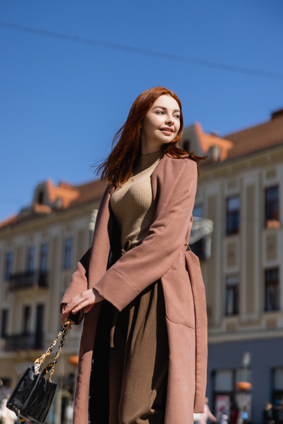 nízký úhel pohledu na usmívající se zrzka žena v kabátu na ulici městského evropského města  - Fotografie, Obrázek