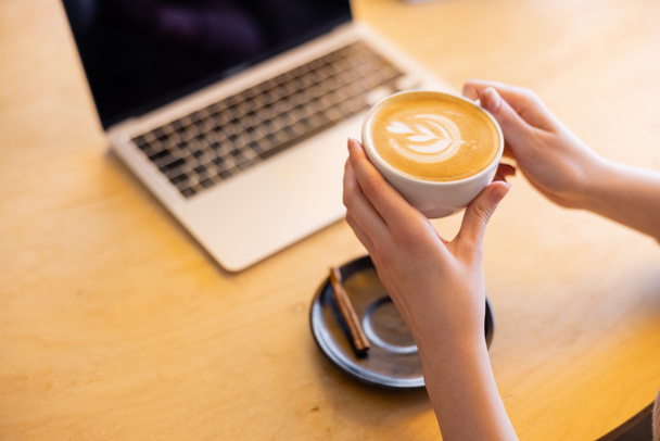 cropped view of young freelancer holding cup of cappuccino near laptop in cafe   - Photo, image