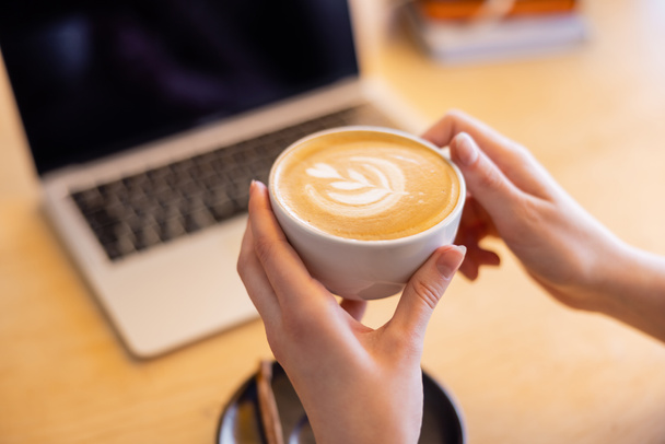 cropped view of young woman holding cup of cappuccino near laptop in cafe   - Фото, изображение