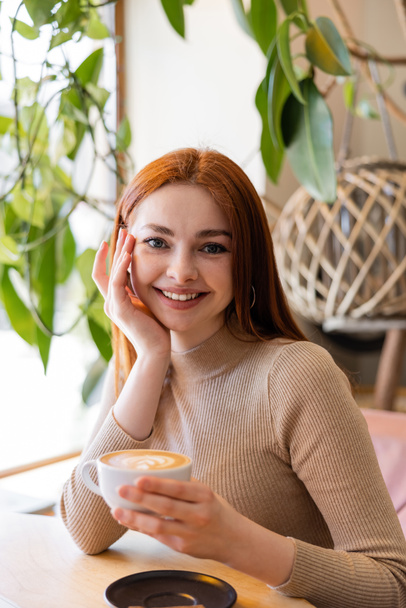 young woman smiling and holding cup of cappuccino in cafe  - Foto, Imagen