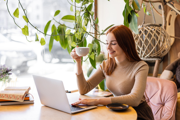 smiling woman with red hair holding cup of coffee and using laptop in cafe  - Фото, изображение