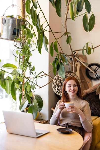 szczęśliwa kobieta z czerwonymi włosami trzyma filiżankę kawy w pobliżu laptopa w kawiarni  - Zdjęcie, obraz