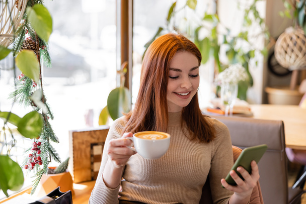 happy young woman with red hair using smartphone and holding cup of coffee in cafe - Photo, image