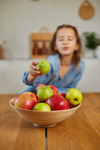 Szczęśliwa dziewczynka bierze gruszkę z miski z różnymi owocami na stole w domu, dziecko jedzące zdrową przekąskę, wegetariańskie odżywianie dla dzieci, witaminy dla dzieci. - Zdjęcie, obraz