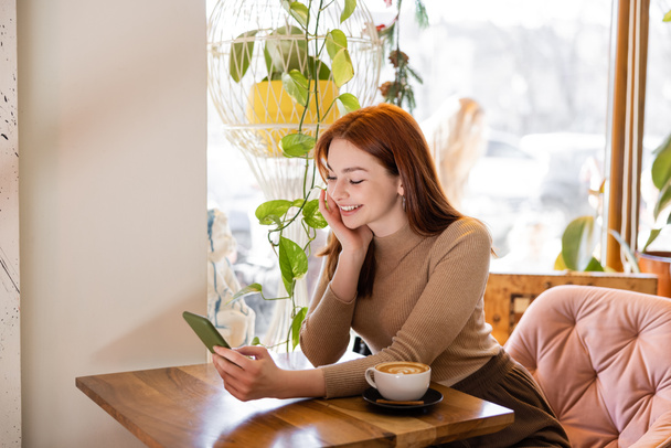 счастливая женщина с рыжими волосами с помощью смартфона возле чашки латте в кафе - Фото, изображение
