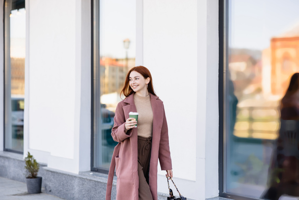 веселая рыжая женщина с сумочкой, держащая кофе, чтобы выйти на городскую улицу  - Фото, изображение