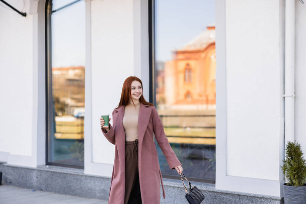 cheerful redhead woman with handbag holding paper cup on street  - 写真・画像