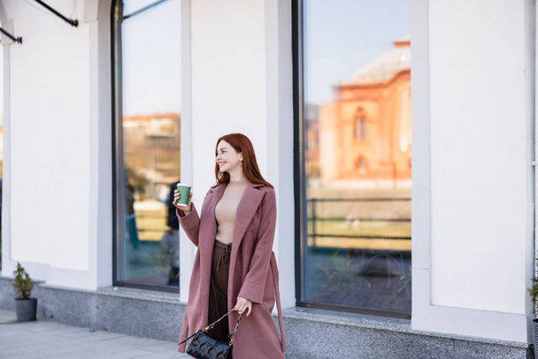 happy redhead woman with handbag holding paper cup on street  - Fotó, kép