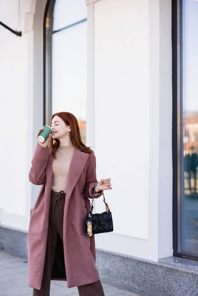 pleased woman with closed eyes drinking coffee to go on urban street  - Fotografie, Obrázek