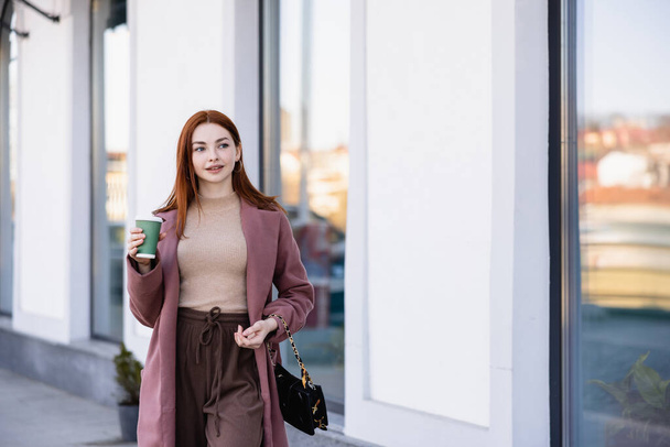 young pleased woman in coat holding coffee to go on street  - Фото, зображення