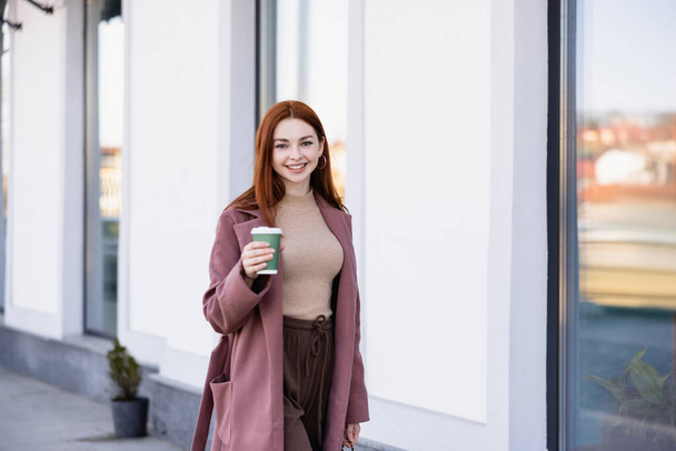 cheerful woman in coat holding coffee to go on urban street  - Fotoğraf, Görsel
