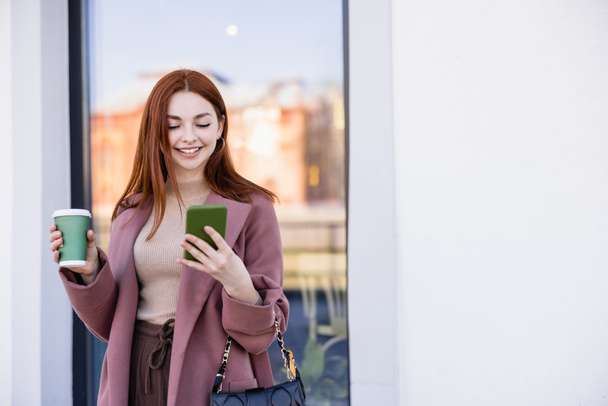 mujer alegre en abrigo sosteniendo café para ir y mirando el teléfono inteligente en la calle urbana - Foto, Imagen