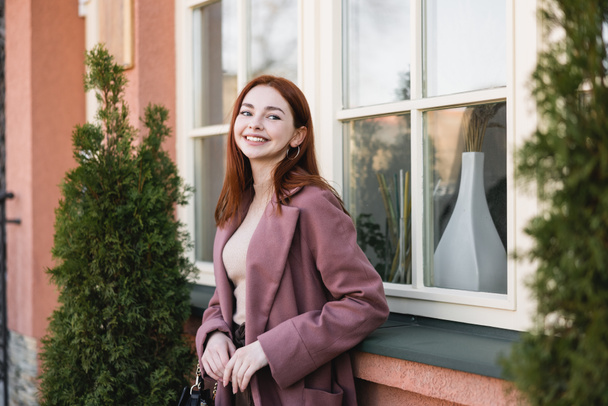 νεαρή χαρούμενη γυναίκα με κόκκινα μαλλιά στέκεται κοντά στο κτίριο  - Φωτογραφία, εικόνα