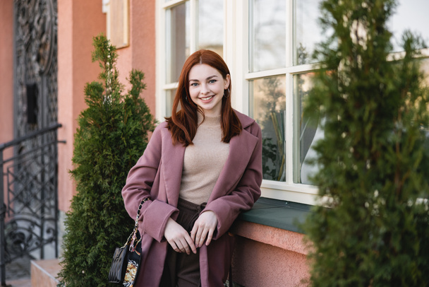 junge fröhliche Frau mit roten Haaren steht in der Nähe des Gebäudes  - Foto, Bild