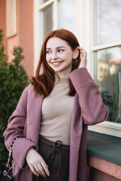 young pleased woman adjusting red hair near building - Photo, Image