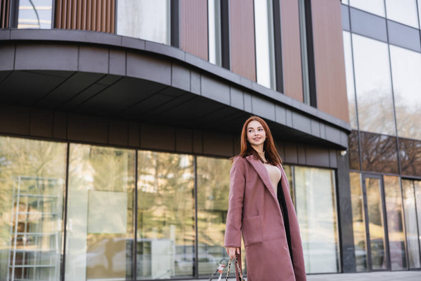 pleased redhead woman in coat standing near modern building  - 写真・画像