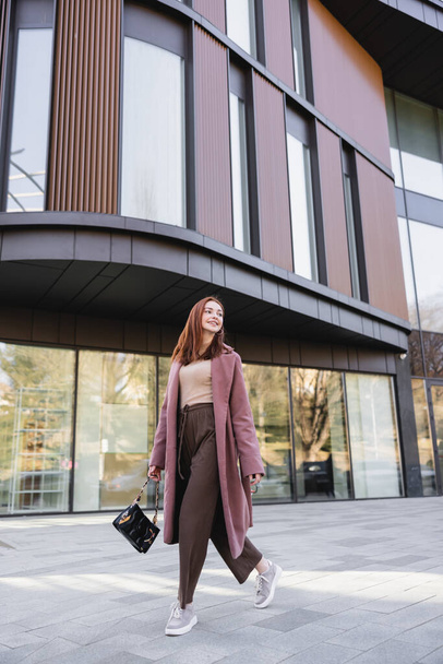 teljes hossza elégedett vörös hajú nő kabátban kézitáskával séta közel modern épület  - Fotó, kép