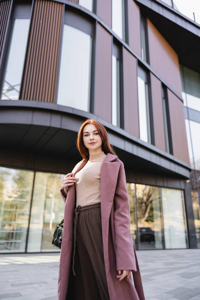 low angle view of redhead woman in coat standing with handbag near modern building  - Photo, image