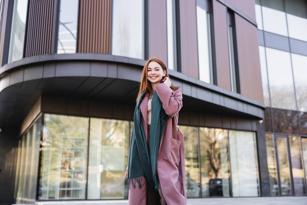 pleased redhead woman in coat with scarf near modern building  - Foto, Bild