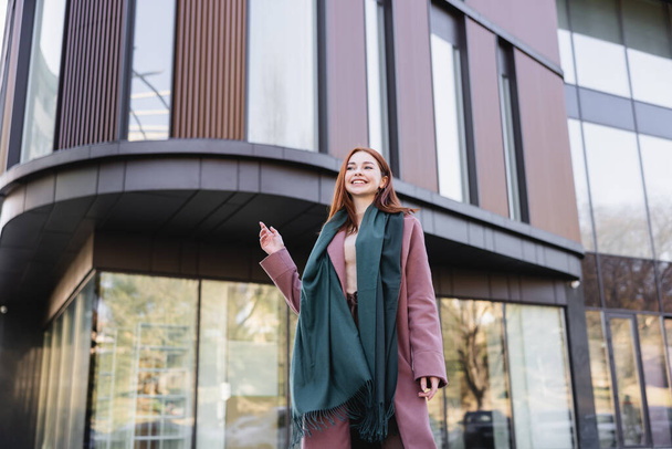 low angle view of joyful redhead woman in coat with scarf near modern building  - Foto, Imagen