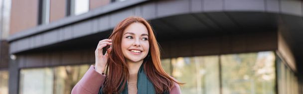 young pleased woman in scarf adjusting red hair near modern building, banner - Foto, immagini