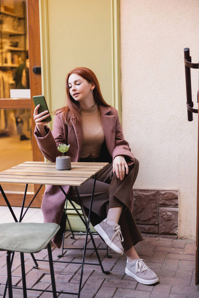 полная длина молодой женщины с рыжими волосами с помощью смартфона на летней террасе  - Фото, изображение