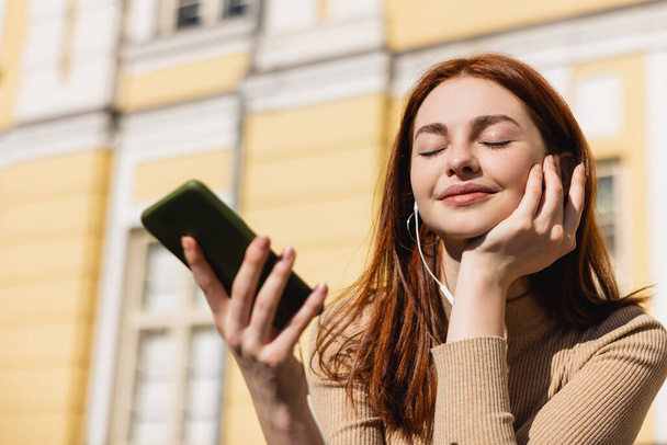 cheerful woman listening music in wired earphones and holding smartphone  - Foto, afbeelding