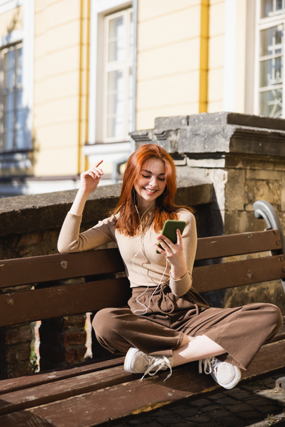 pleased woman listening music in wired earphones and holding smartphone while sitting on bench - Foto, imagen