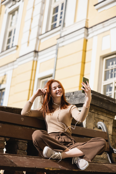 pleased woman listening music in wired earphones taking selfie while sitting on bench - Fotoğraf, Görsel