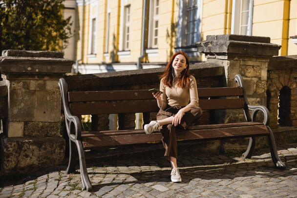 full length of pleased woman listening music in wired earphones and holding smartphone while sitting on bench - Fotoğraf, Görsel