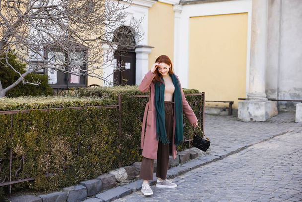 πλήρες μήκος του χαρούμενα νεαρή γυναίκα σε παλτό και μαντήλι κρατώντας τσάντα και το περπάτημα στο δρόμο  - Φωτογραφία, εικόνα
