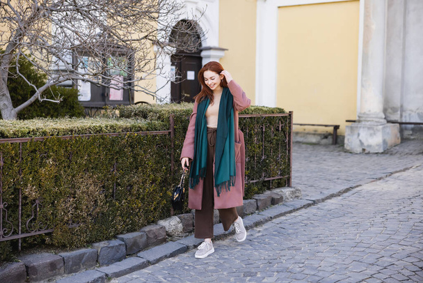 full length of positive young woman in coat and scarf holding handbag and walking on street  - Zdjęcie, obraz