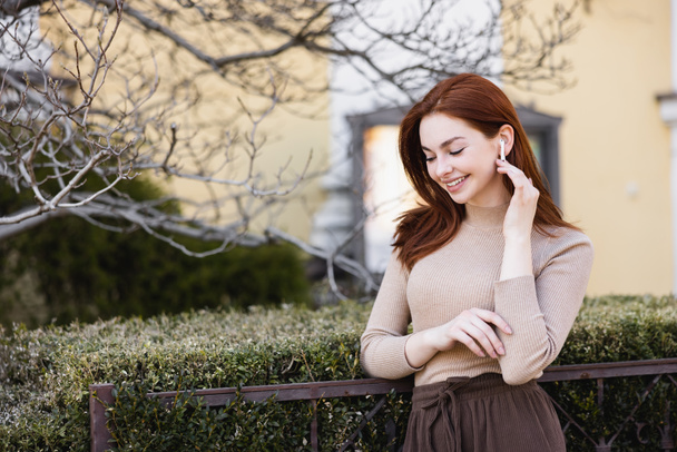 happy and redhead woman adjusting wireless earphone outside - Photo, Image