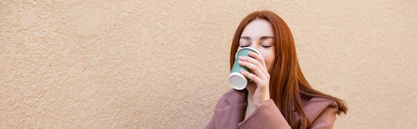 mladá zrzka žena se zavřenýma očima pije kávu jít, prapor - Fotografie, Obrázek