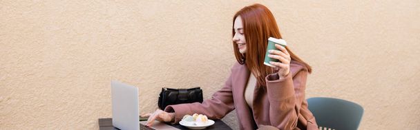šťastný freelancer drží kávu jít a pomocí notebooku v blízkosti desky s macarons, banner - Fotografie, Obrázek