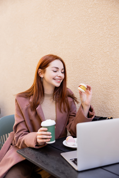 cheerful woman holding coffee to go and macaron near laptop on cafe terrace  - Fotoğraf, Görsel