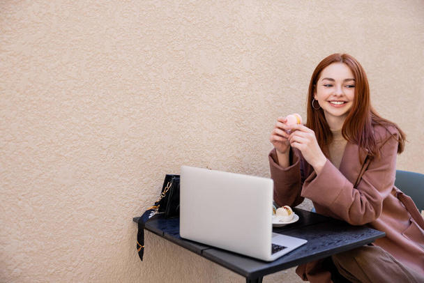 freudige junge Frau mit roten Haaren hält süßen Makkaron in der Nähe von Laptop auf Café-Terrasse - Foto, Bild