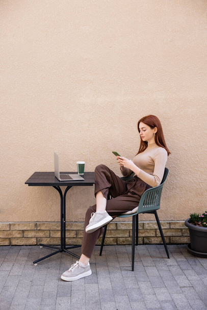 full length of redhead woman in turtleneck using smartphone near laptop on summer terrace  - 写真・画像