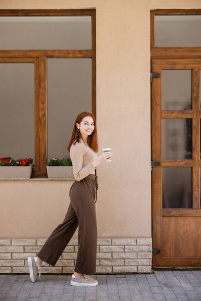 full length of happy young woman in wireless earphone holding paper cup and walking on street - Photo, Image