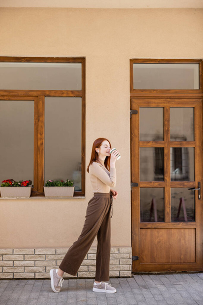 full length of stylish redhead woman in earphone drinking coffee to go  - Foto, Imagem