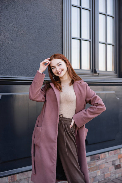 cheerful redhead woman in stylish coat standing with hand on hip near modern building  - Foto, Imagen
