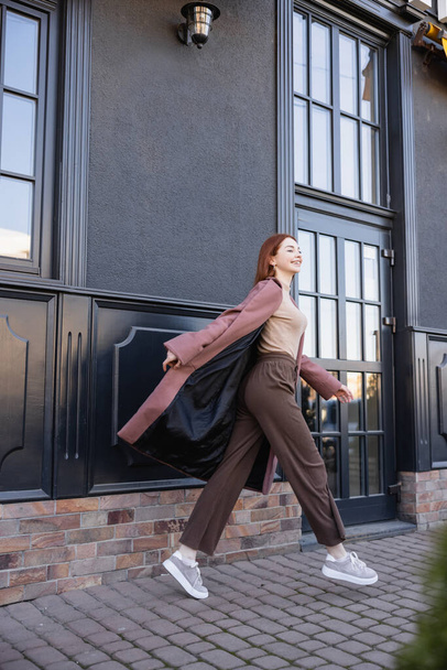 longitud completa de mujer pelirroja alegre en abrigo elegante caminando cerca de edificio moderno  - Foto, Imagen